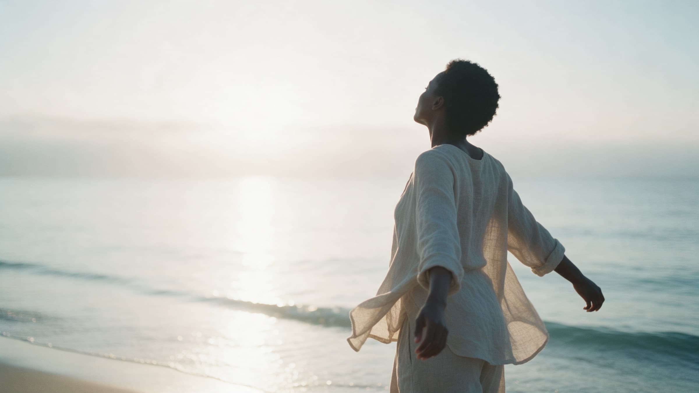 A woman stands at the water's edge at dawn, arms open, face turned to the light.