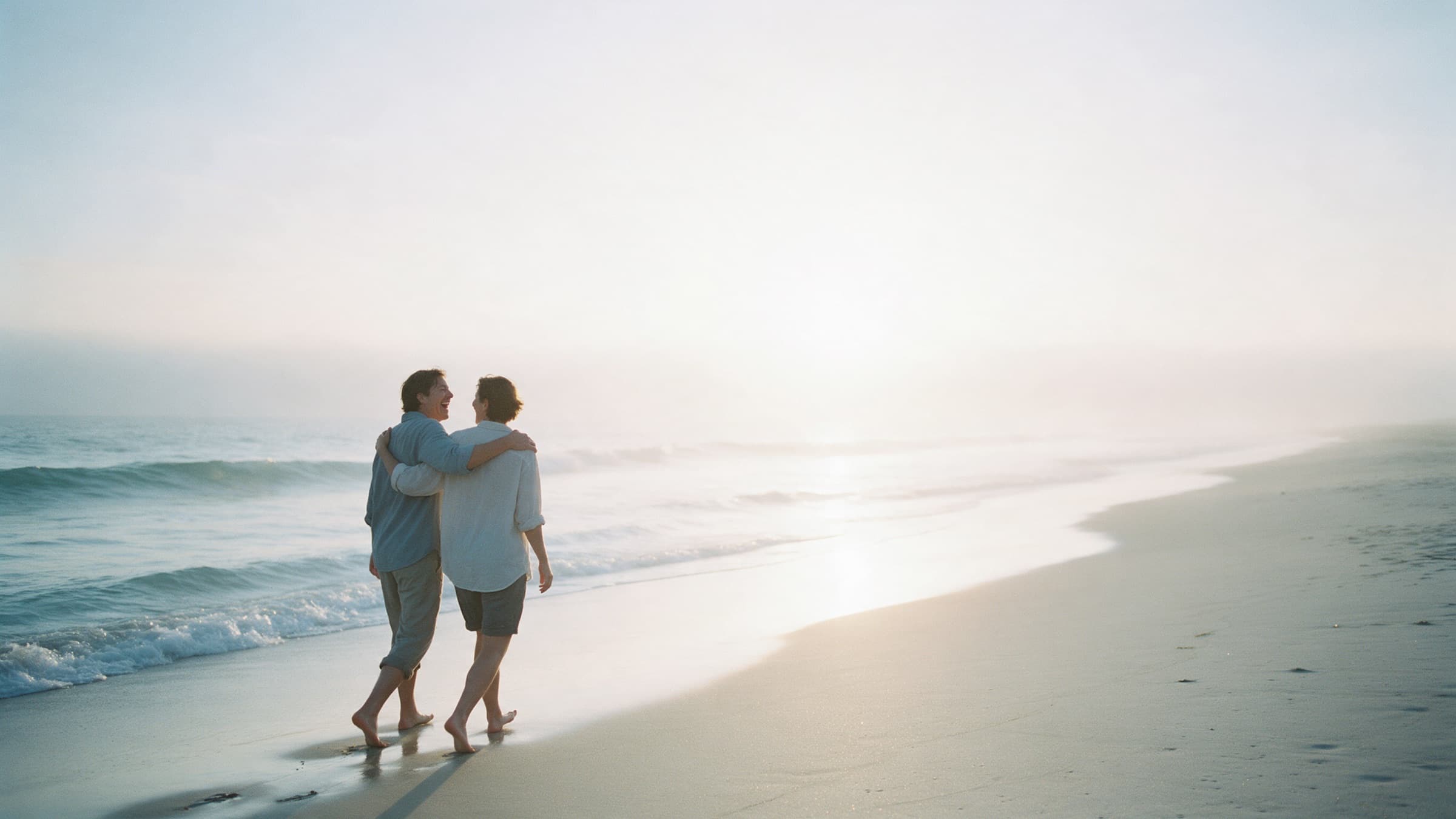 Two friends walk arm in arm along the shoreline in the morning light.